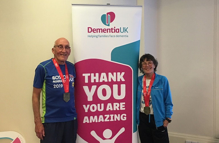Husband and wife standing next to Dementia UK sign, wearing medals and smiling