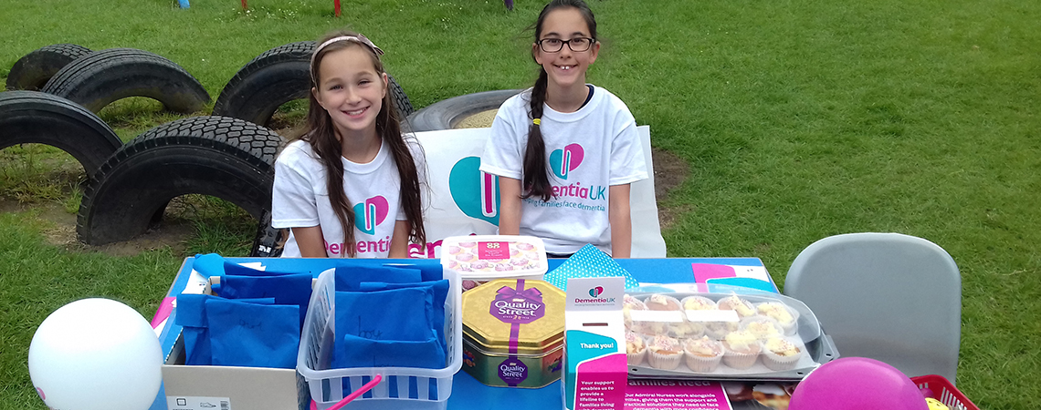 Two young girls wearing Dementia UK t-shirts holding a bake sale outdoors