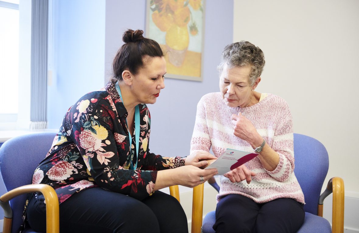 An Admiral Nurse showing a leaflet to a elderly lady. Both of them are sat down.