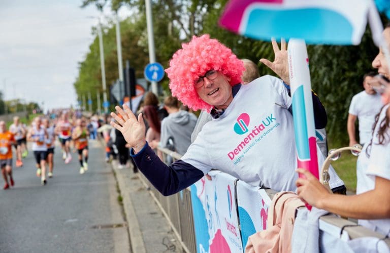 A man in a clown wig at the side of the road at a Dementia UK running event.