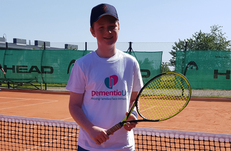 Young Alfie on the tennis court holding a racket and wearing a Dementia UK t-shirt