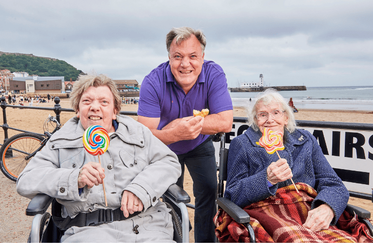 Ed Balls at the seaside eating a doughnut with two older ladies with lollies.