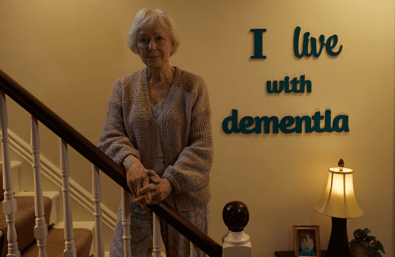 elderly lady standing on the stairs with the lettering I live with dementia on the wall behind her