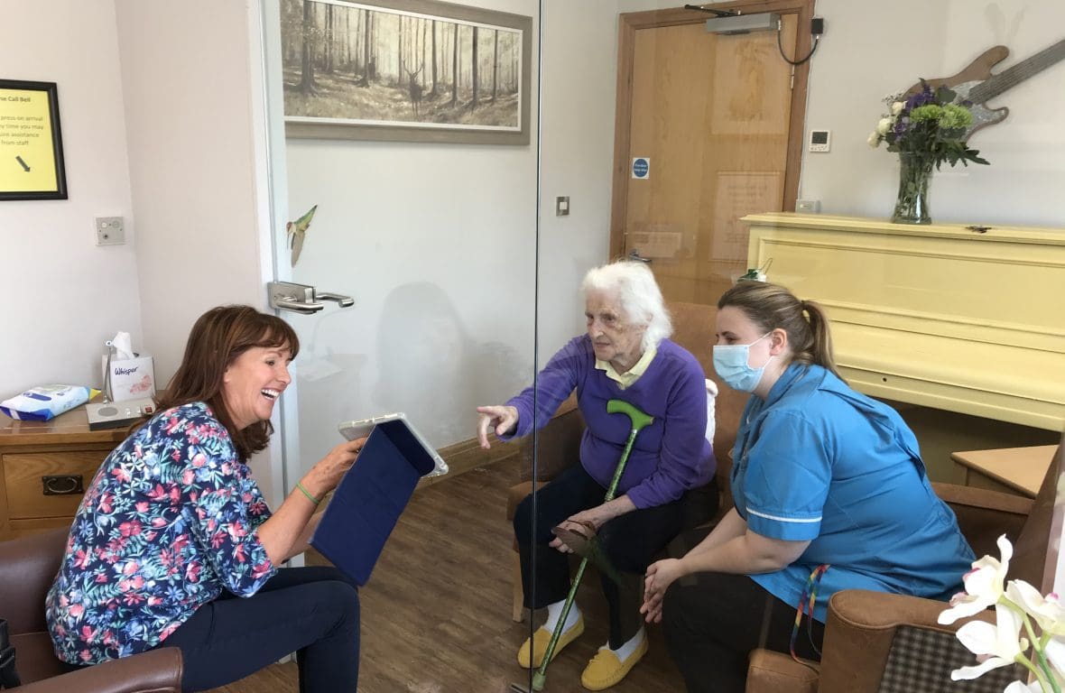 woman showing pictures to elderly female relative with a glass divider between them. A carer is sat next to the elderly women.