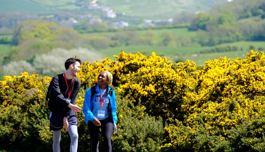 Man and woman talking and laughing together while walking. Behind them is a countryside view and hedges of flowers