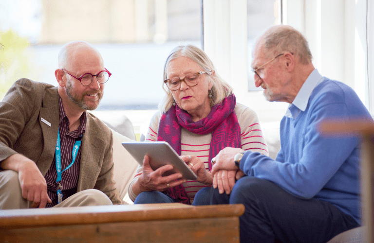 elderly man and woman are sitting at a coffee table reviewing information on an ipad, shown to them by a male admiral nurse