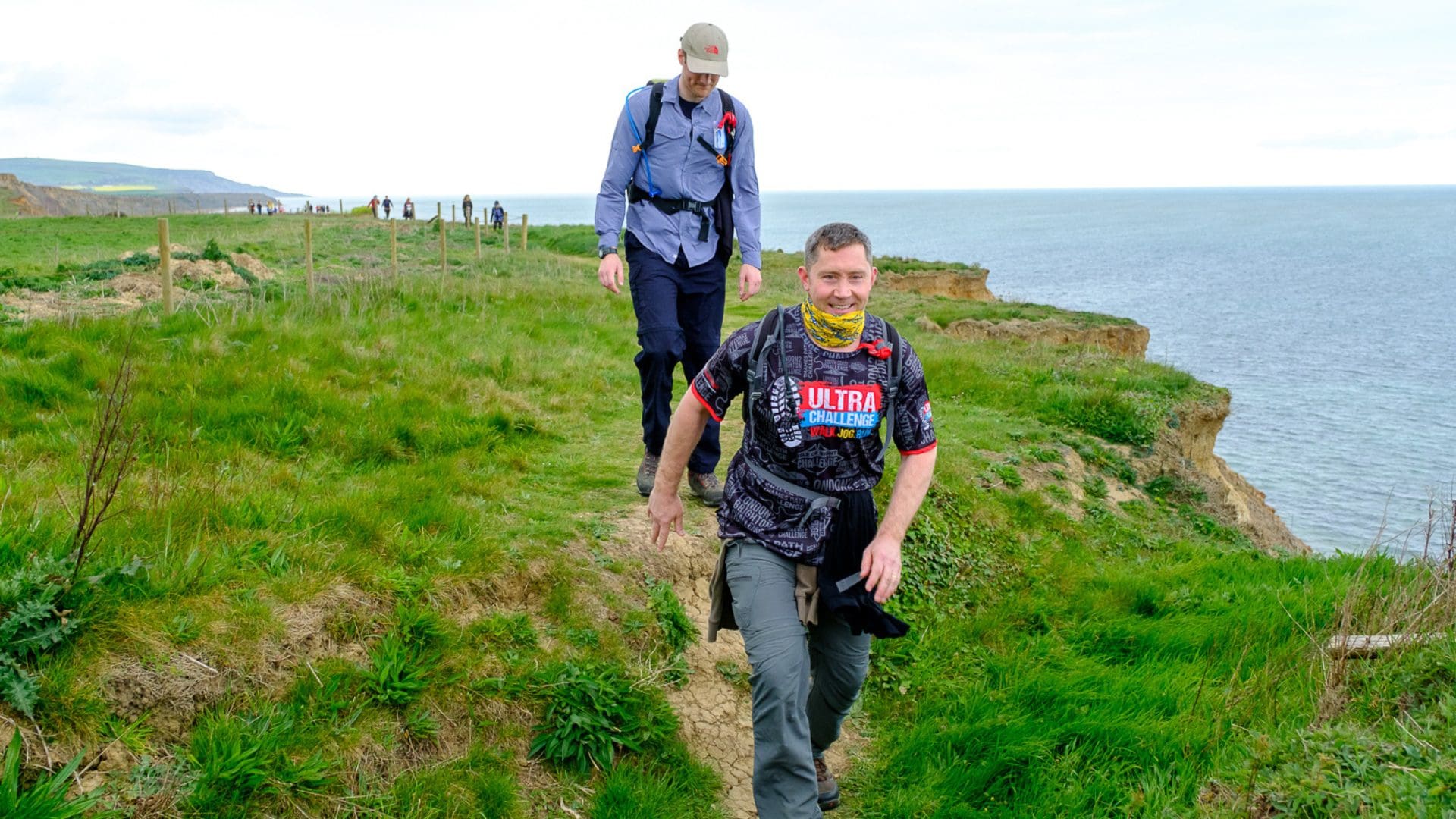 Two men walking towards us on the cliff top with the coastline next to them