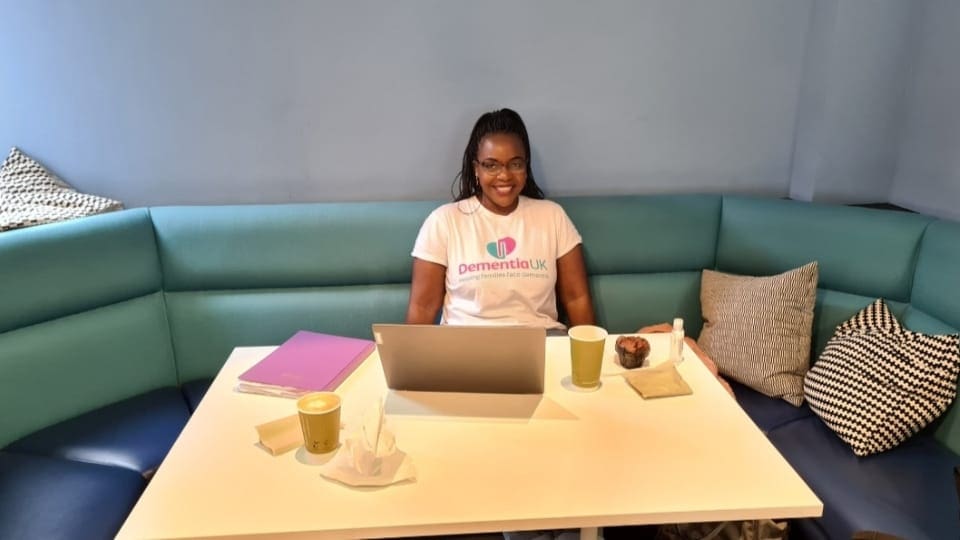 woman seated with laptop on the desk in front of them. They are smiling and are wearing a Dementia UK branded tee shirt