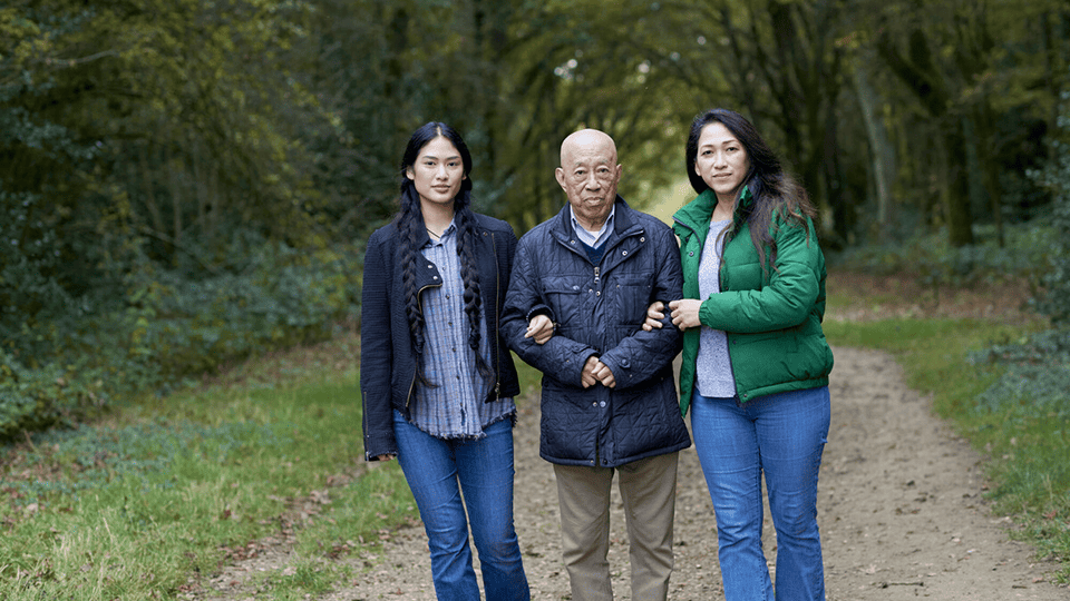 A young woman accompanies and older woman and much older man on a walk through a green parkland.