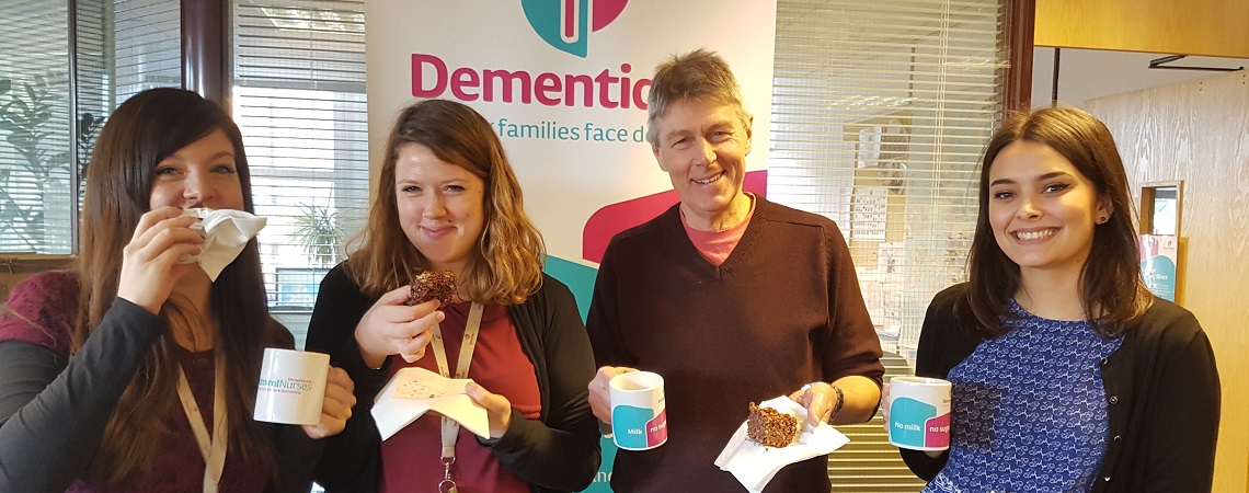 Four Dementia UK staff members standing in front of a Dementia UK sign sipping tea and eating cake