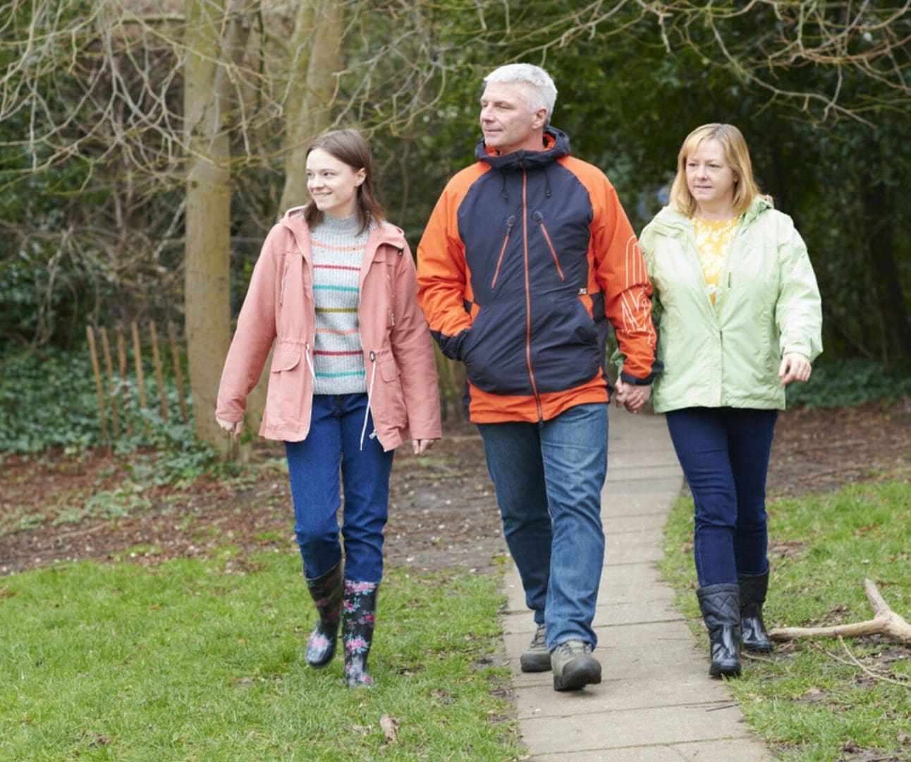 A family of three are walking down a pathway through a park holding hands. From the left there is a daughter, father, and mother