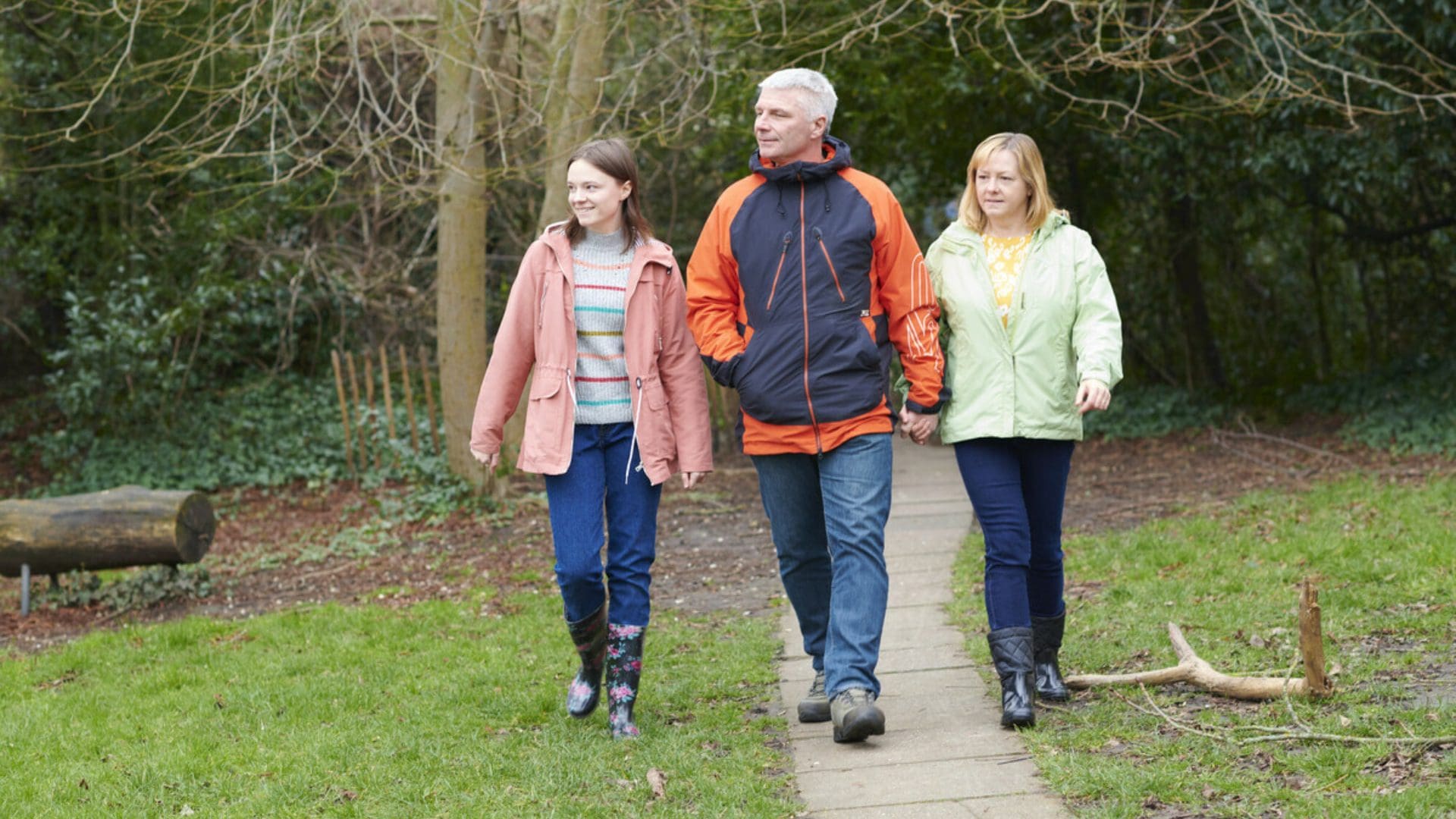 A family of three are walking down a pathway through a park holding hands. From the left there is a daughter, father, and mother