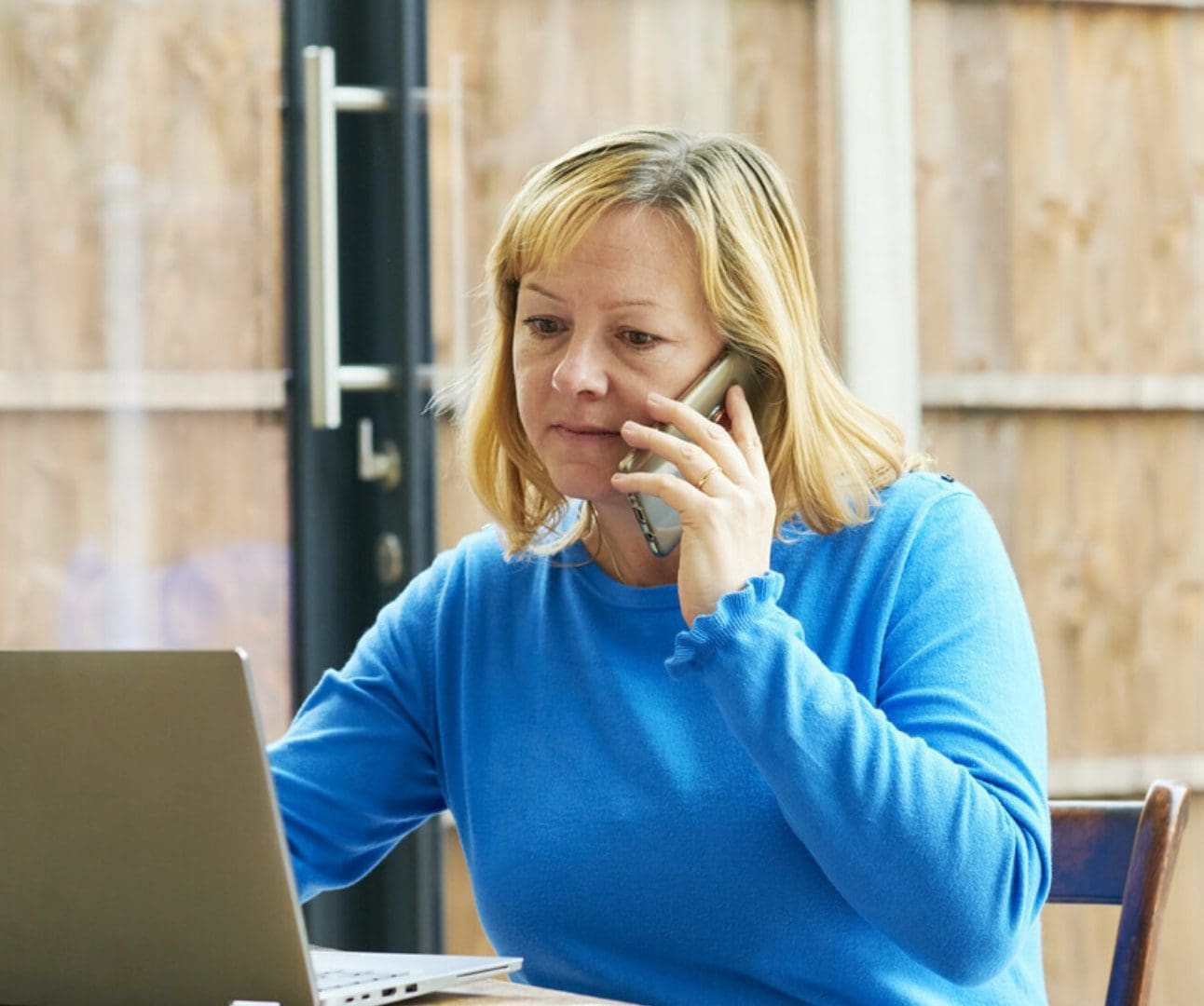 woman sitting at a table looking at her open laptop whilst making a phone call
