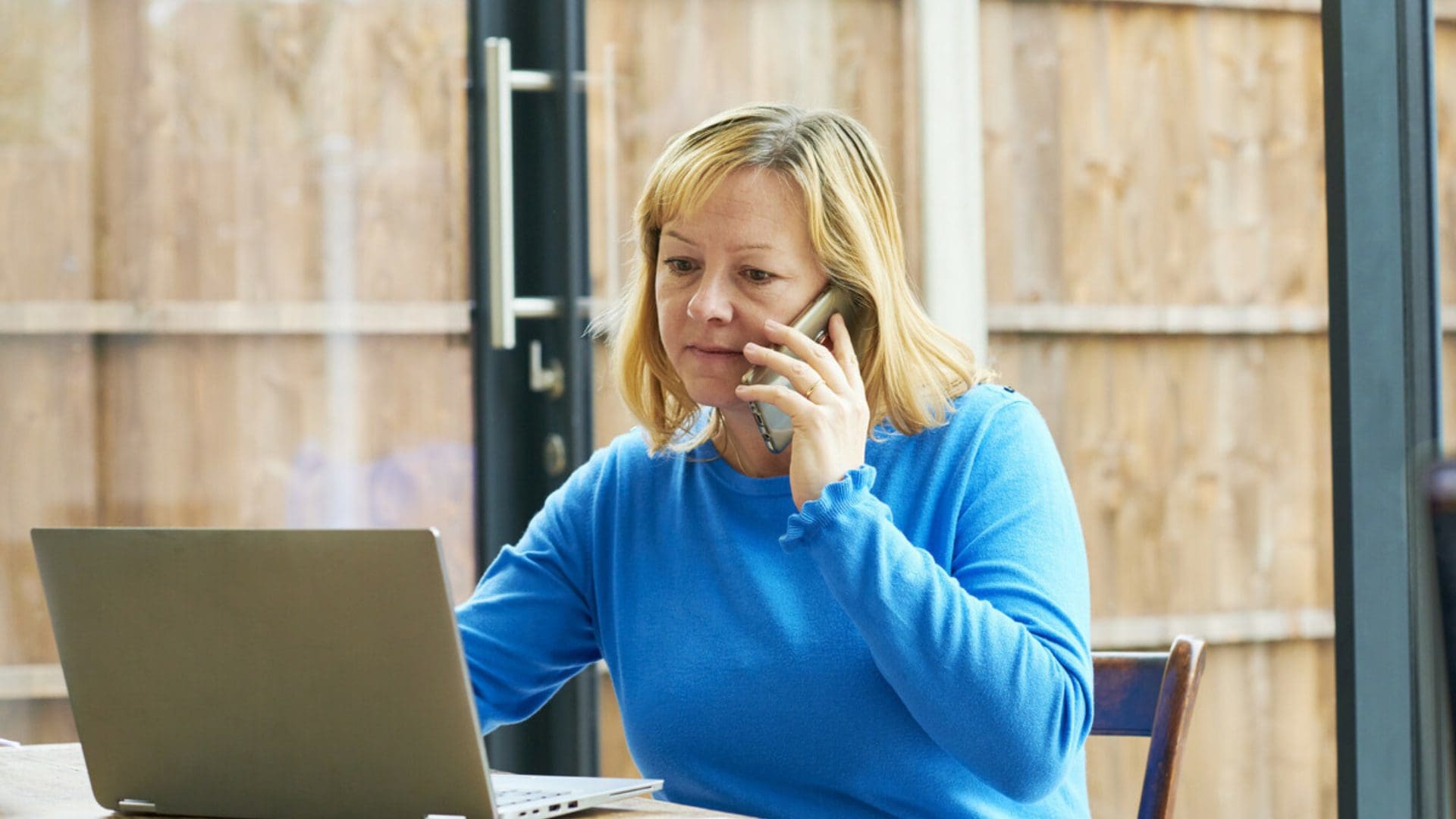 woman sitting at a table looking at her open laptop whilst making a phone call