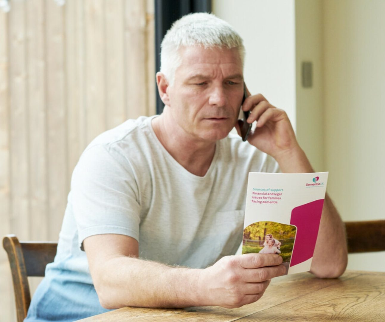 man sat at the dining table on the phone whilst looking at a Dementia UK leaflet