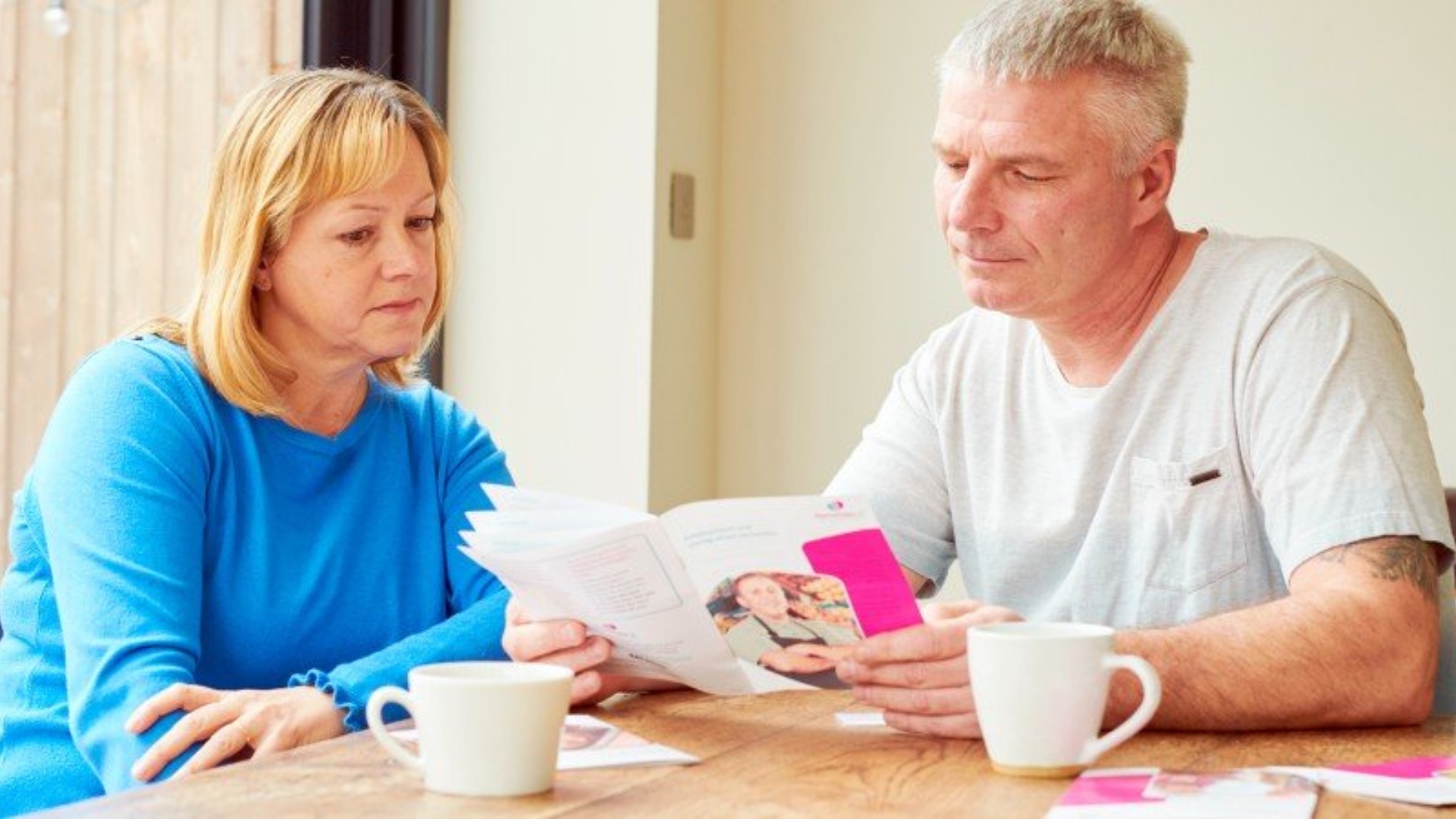 Husband and wife sat at the dining table reading a Dementia UK leaflet with two mugs of tea